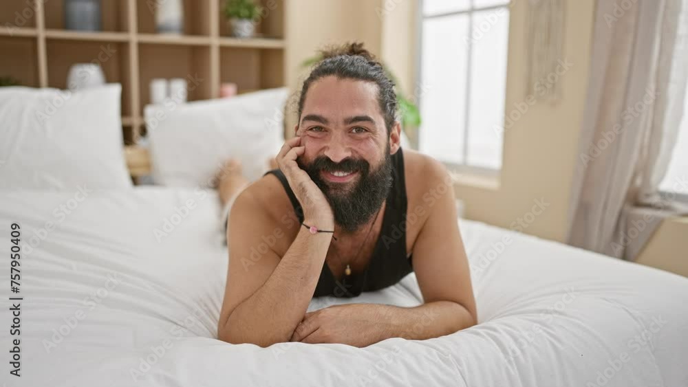 Handsome hispanic man with a beard smiling while lying on a white bed in a bright bedroom indoors Handsome hispanic man with a beard smiling while lying on a white bed in a bright bedroom indoors