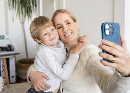 Smiling young mother and little son using phone together, hugging, at home, happy mum with adorable kid boy taking selfie, enjoying leisure time with gadget