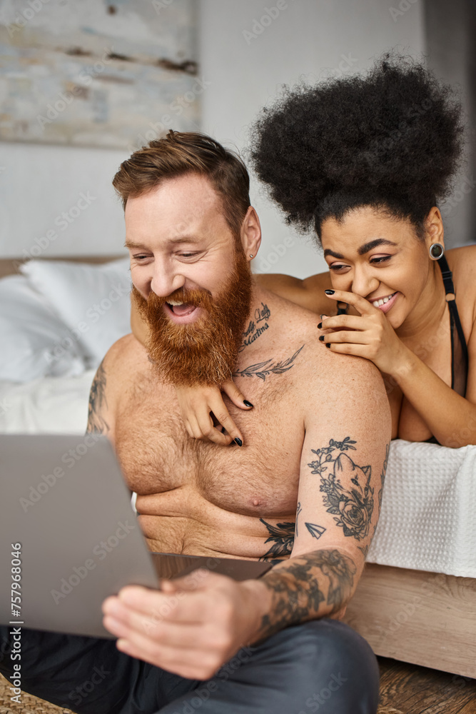 Fototapeta premium happy tattooed man and african american woman watching comedy movie on laptop, multicultural couple