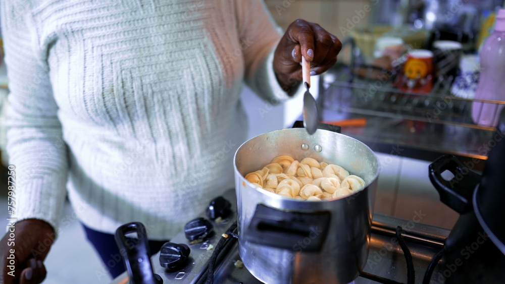 Fototapeta premium One elderly South American black woman standing in kitchen stove cooking pasta. Gray Haired 80s person of African descent preparing lunch