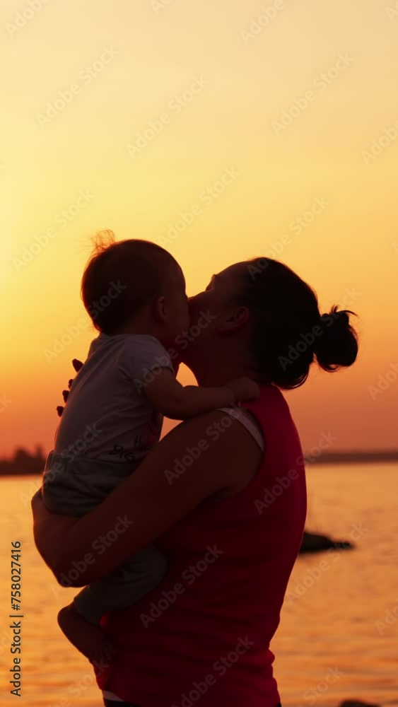 Brunette woman in red sleeveless shirt holding a little toddler. Mother and son enjoying beautiful sunset near the river. Vertical video. Brunette woman in red sleeveless shirt holding a little toddler. Mother and son enjoying beautiful sunset near the river. Vertical video.