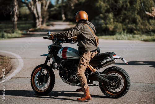 Chico con barba con atuendo de motorista posando con su moto y su cámara 