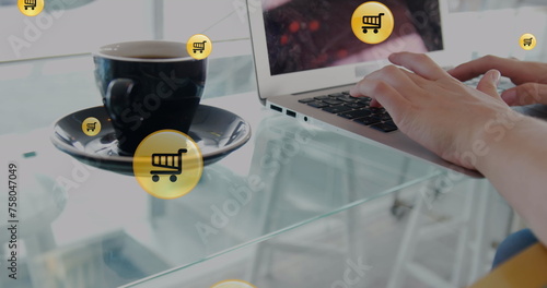 Image of shopping cart icons, coffee cup, cropped hands of caucasian woman working on laptop