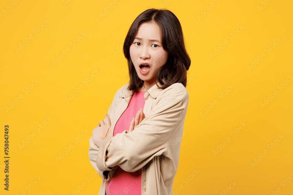 Side view young frowning sad woman of Asian ethnicity she wears pink t-shirt beige shirt pastel casual clothes hold hands crossed folded isolated on plain yellow background studio. Lifestyle portrait. Side view young frowning sad woman of Asian ethnicity she wears pink t-shirt beige shirt pastel casual clothes hold hands crossed folded isolated on plain yellow background studio. Lifestyle portrait.