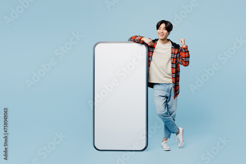 Full body young man of Asian ethnicity wear red hoody casual clothes big huge blank screen mobile cell phone smartphone with area do winner gesture isolated on plain pastel light blue cyan background