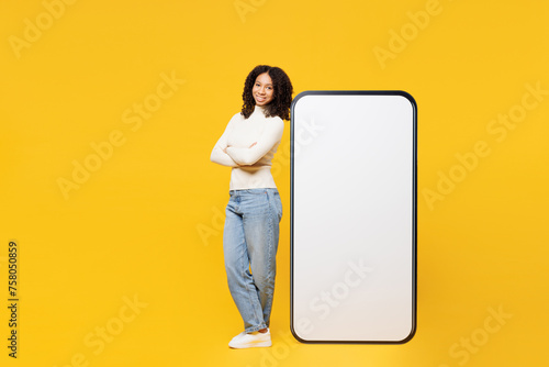 Full body fun little kid teen girl wear white casual clothes stand near big huge blank screen mobile cell phone smartphone with workspace area isolated on plain yellow background. Lifestyle concept. photo