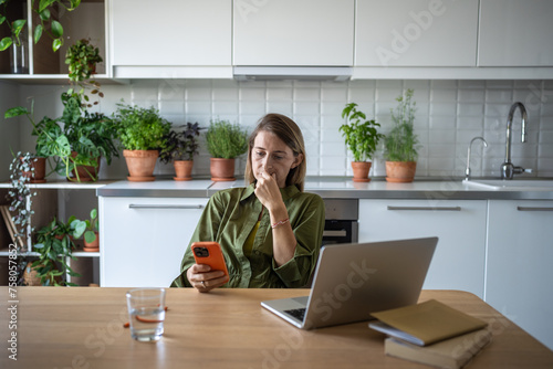 Photography Saddened stressed female sitting at home table with laptop reading disturbing news on mobile phone