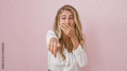 Joyful young blonde woman caught in a cheerful prank! standing in a pink isolated background, wearing a shirt, pointing finger at you, laughing with hand over mouth. can you match her happiness?