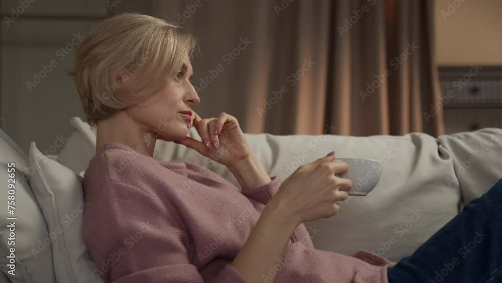 A contemplative mature woman is seated comfortably on a white sofa, holding a cup of tea in her hand. He gazes thoughtfully into the distance, perhaps unwinding after a long way or indulging in a A contemplative mature woman is seated comfortably on a white sofa, holding a cup of tea in her hand. He gazes thoughtfully into the distance, perhaps unwinding after a long way or indulging in a