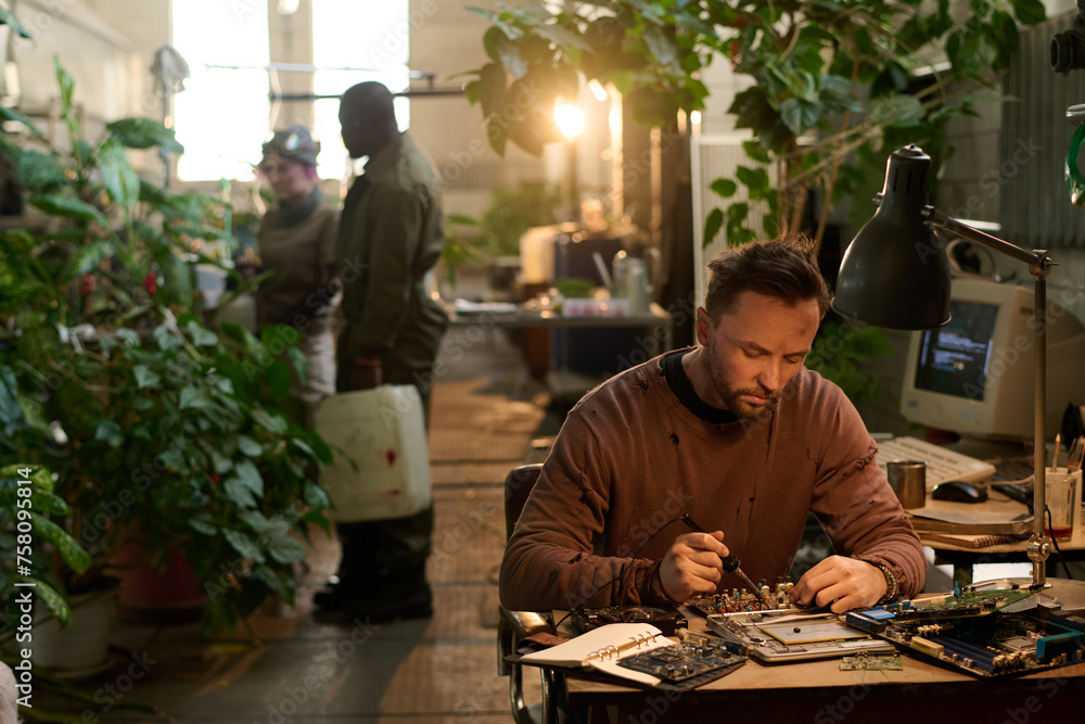Selective focus shot of Caucasian man repairing old computer while his friends taking care of plants in post-apocalyptic shelter Selective focus shot of Caucasian man repairing old computer while his friends taking care of plants in post-apocalyptic shelter