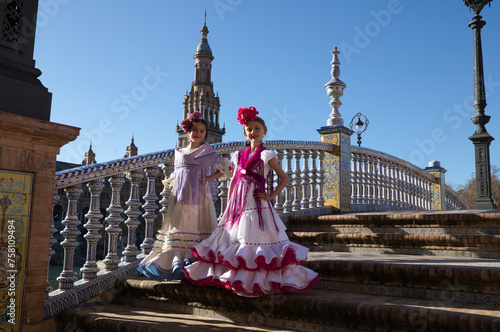 Two pretty little girls dancing flamenco dressed in typical gypsy costume pose on the stairs in their dress in a famous square in seville. The girls are happy. Flamenco, cultural heritage of humanity.
