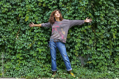 A young model beautiful woman girl in denim trousers stands near a wall overgrown with climbing plants. Background with plant leaves.