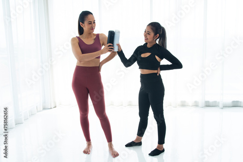 Confident woman in black sportswear presenting a water bottle to her friend in maroon activewear against a bright, minimalist backdrop © Arunporn