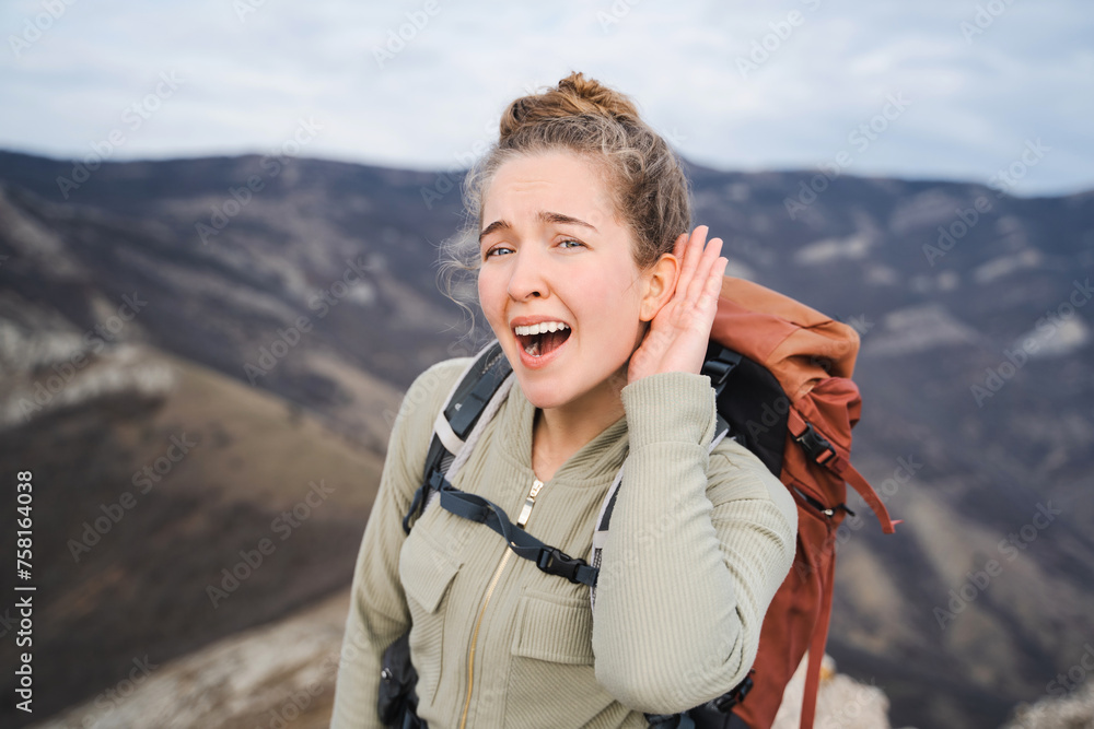 What? I can't hear you! Young female hiker with tourist backpack holding hand near ear and listening carefully, keeps mouth opened, having hearing problems outdoor, deafness in 