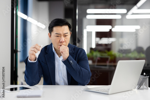 Young Asian male businessman sitting at desk in office and suffering from severe cough and asthma attack, holding napkin and covering mouth with hand.