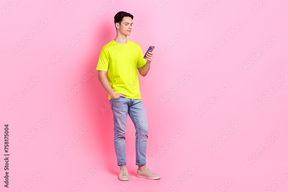 Full length photo of cheerful good mood guy wear green t-shirt texting modern device empty space isolated pink color background Full length photo of cheerful good mood guy wear green t-shirt texting modern device empty space isolated pink color background