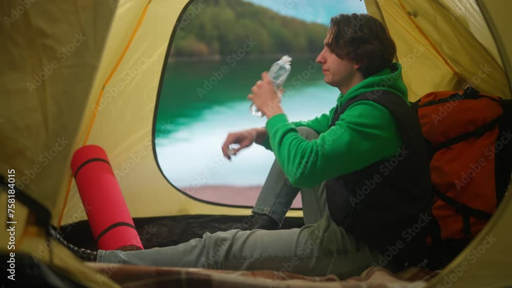 Person on campsite traveling and hiking, exploring nature. Young man sitting drinking water inside the tent at lake coast in mountains, holiday and vacation outdoors. Person on campsite traveling and hiking, exploring nature. Young man sitting drinking water inside the tent at lake coast in mountains, holiday and vacation outdoors.