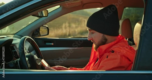 A young man dressed in an orange jacket and cap, in a car with a phone in his hands.