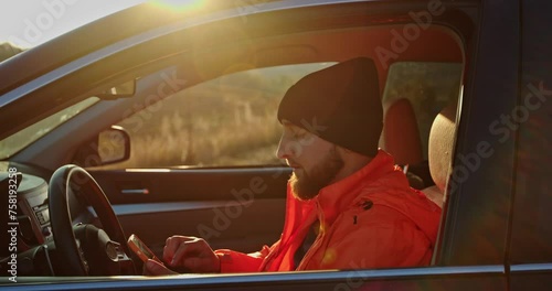 A young man dressed in an orange jacket and cap, in a car with a phone in his hands.