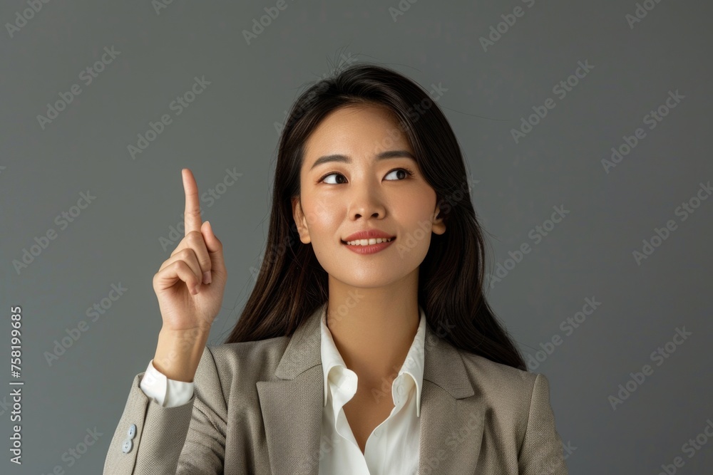 An Asian businesswoman pointing upwards, looking at the camera with a smile, which could indicate optimism or sharing a new idea