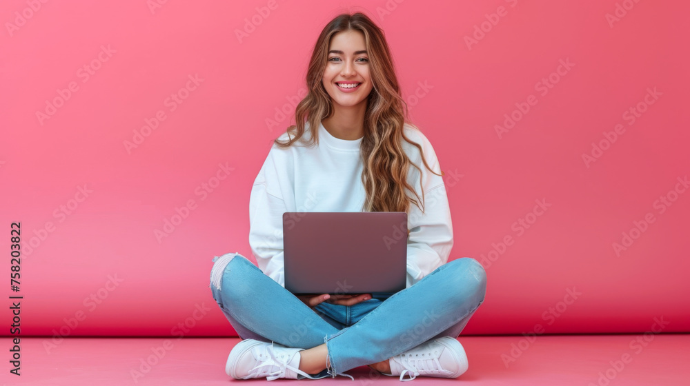 cheerful young woman sitting with her legs crossed, holding a laptop on her lap, wearing a white sweatshirt and ripped blue jeans, against a coral pink background cheerful young woman sitting with her legs crossed, holding a laptop on her lap, wearing a white sweatshirt and ripped blue jeans, against a coral pink background