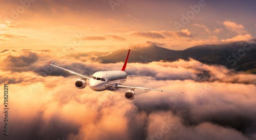 Plane is flying over mountain peaks in low clouds at golden sunrise. Top view of passenger airplane, colorful sky, hills in fog. Aircraft is taking off. Business travel. Commercial. Aerial view