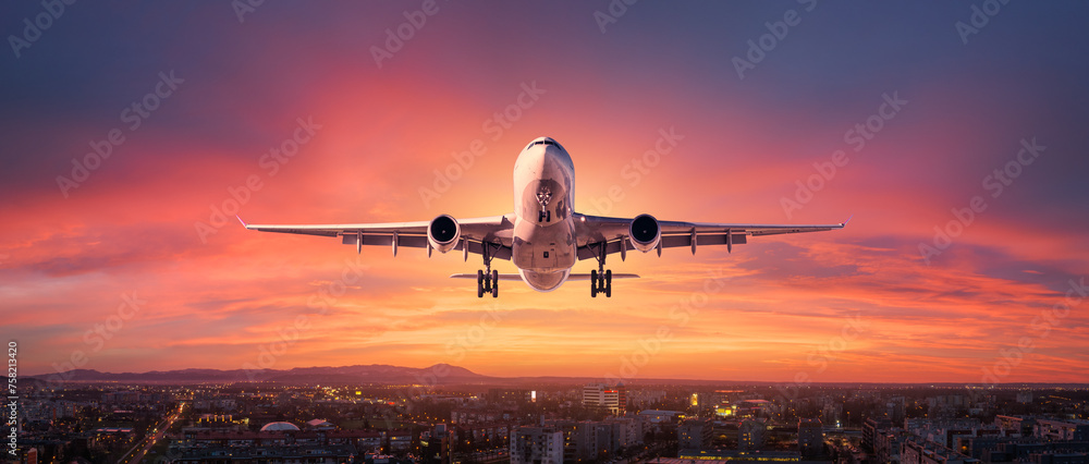 Airplane is flying in colorful sky over city at sunset. Landscape with passenger airplane, skyline, purple sky with red and pink clouds at dusk. Aircraft is landing at twilight. Aerial view of plane - obrazy, fototapety, plakaty 