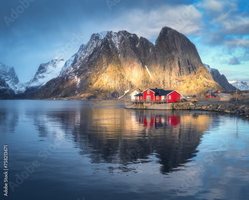 Red rorbuer on the island against snowy rocks lighted by sun, blue sea and dramatic cloudy sky in winter. Landscape with houses, mountains at sunset. Rorbu in Hamnoy village, Lofoten islands, Norway