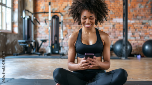 young woman is seated on a gym floor in workout attire, looking at her smartphone, with weightlifting equipment in the background
