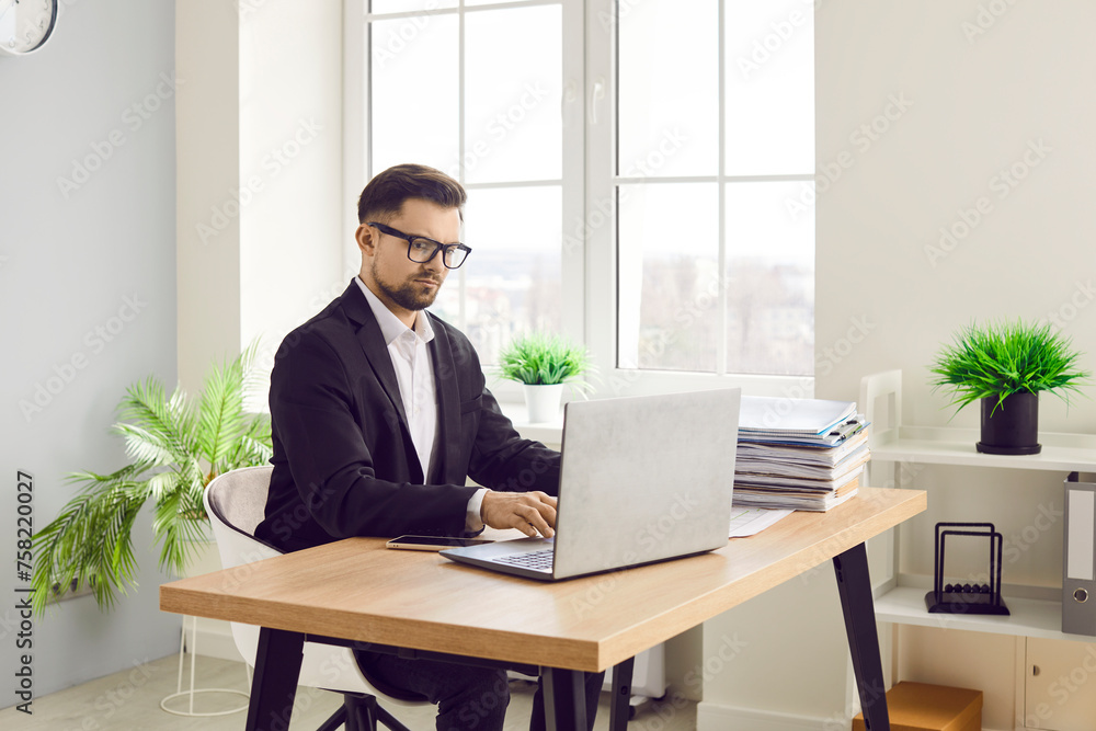 Focused confident businessman working with laptop in office. Portrait of handsome bearded man in formal wear and eyeglasses sitting at desk and typing on laptop computer Focused confident businessman working with laptop in office. Portrait of handsome bearded man in formal wear and eyeglasses sitting at desk and typing on laptop computer