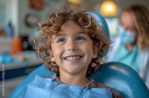 Cheerful boy with curly hair sits in a dental chair, with a blurred dentist in the background