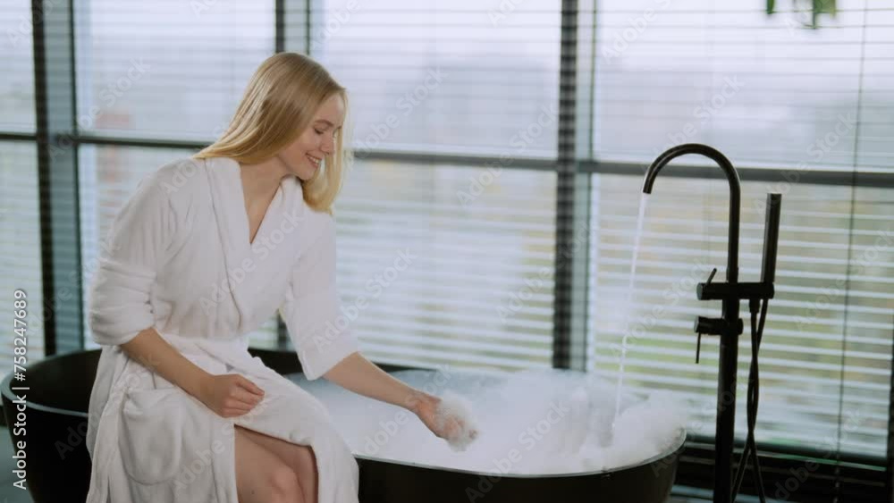 Girl preparing bath with foam to wash. Relaxed positive woman in bathrobe waiting throwing soapy foam cloud in air and catching it by hands in bathroom. Home spa wellness self care leisure concept. Girl preparing bath with foam to wash. Relaxed positive woman in bathrobe waiting throwing soapy foam cloud in air and catching it by hands in bathroom. Home spa wellness self care leisure concept.