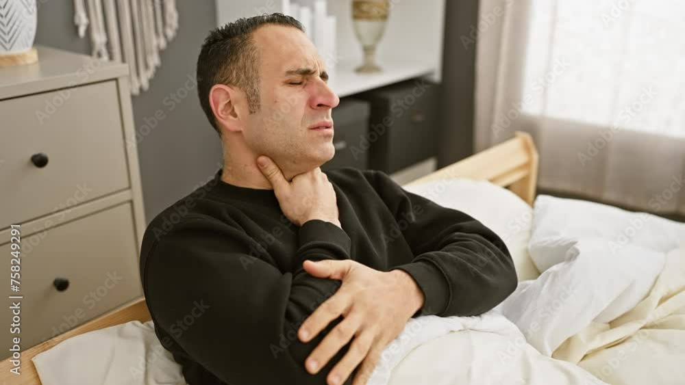 A man in pain holding his throat while sitting in bed in a bedroom displaying symptoms of sore throat or illness, captured for a medical or health-related context. A man in pain holding his throat while sitting in bed in a bedroom displaying symptoms of sore throat or illness, captured for a medical or health-related context.