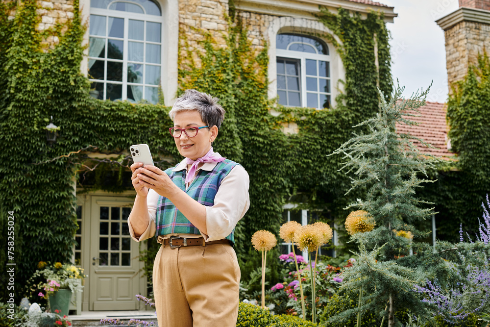 sophisticated joyous mature woman in chic attire looking at her smartphone near her house in England sophisticated joyous mature woman in chic attire looking at her smartphone near her house in England