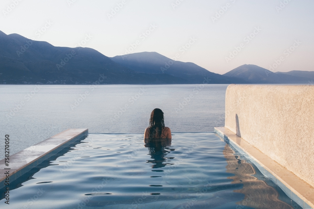 A slim girl with long hair stands with her back in the luxurious infinity outdoor pool with the horizon on the background of the sea and mountains. Woman watching the sunrise. 