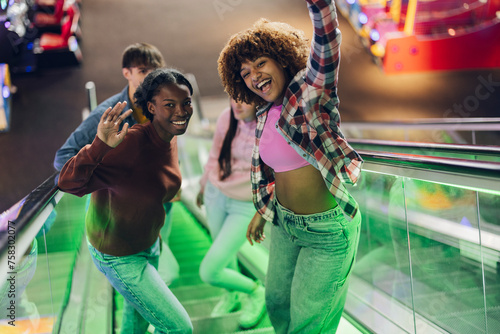 Ethnic friends waving at camera very smiling while climbing escalators with neon lights © Aida