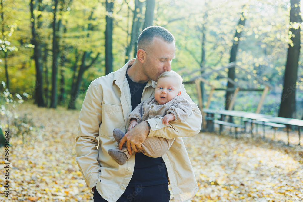 Fototapeta premium Happy young dad hug his little son in the sun on a warm spring or summer day in the park. Sunshine light in the park. Father and son family concept in nature