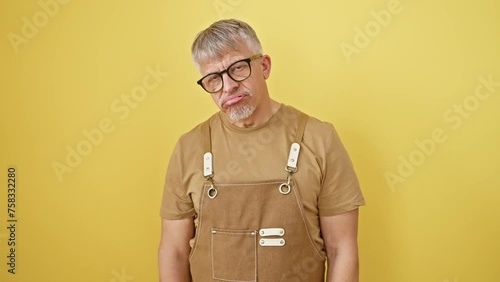 Exhausted middle age man in glasses and apron, looking sleepily at the morning, showing signs of fatigue and hangover, lazy eyes staring from a yellow isolated background photo