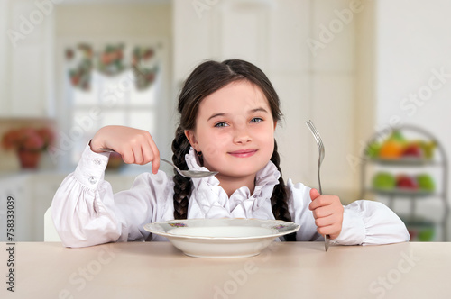 little teenage girl sitting at the dining table in the kitchen. Before appetite eating with a plate, spoon. 