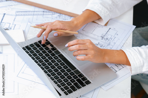 Professional beautiful engineer using laptop analysis blueprint with architectural document and house model placed on table. Creative business design and teamwork concept. Focus on hand. Immaculate.