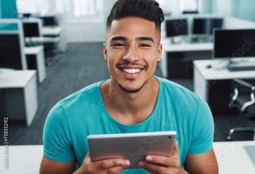 A smiling man in a turquoise t-shirt uses a tablet in an office setting, suggesting a relaxed yet productive environment.