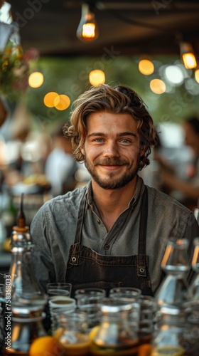 Smiling person with tousled hair, wearing an apron, stands before a table with glass bottles