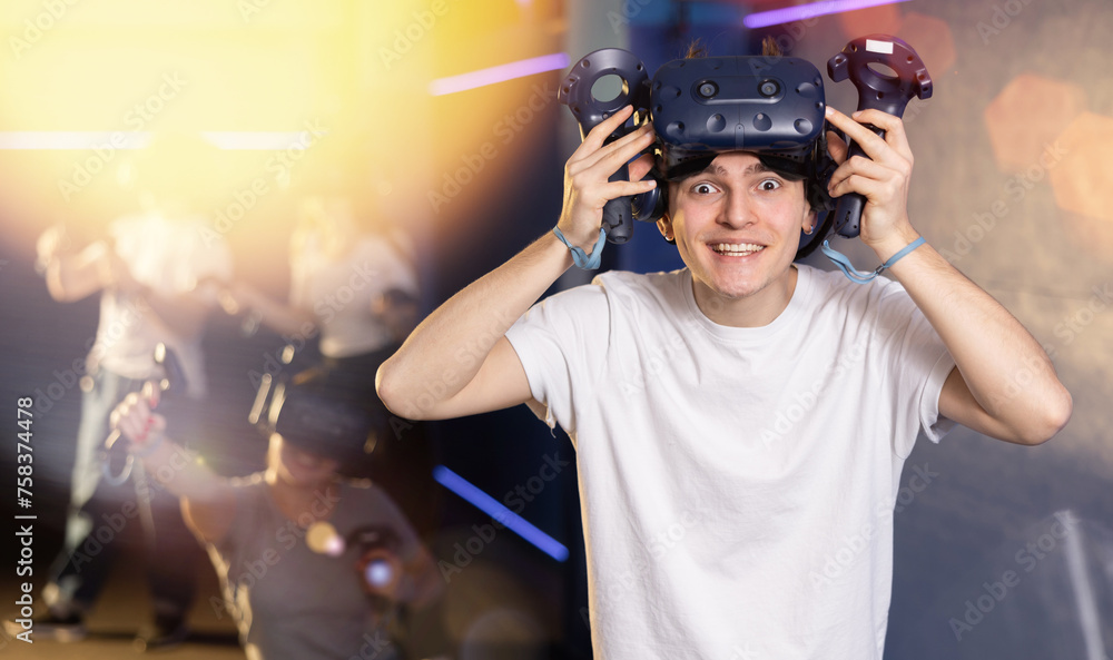 Portrait of happy excited young guy with VR glasses on head, holding handheld controllers playing virtual reality game with friends..
