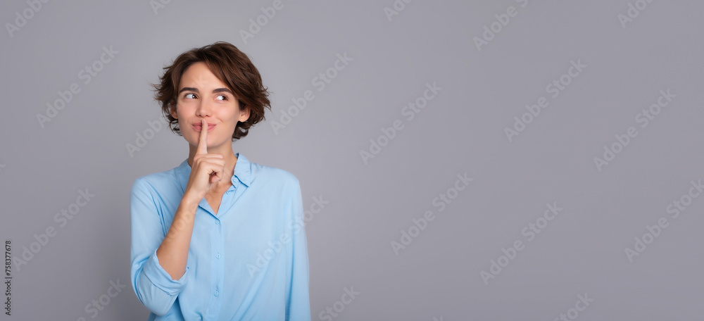 Banner photo of beautiful happy brunette woman wearing smart casual clothes posing saying shush be quiet with finger on her lips shhh gesture looking away isolated on gray wall in studio