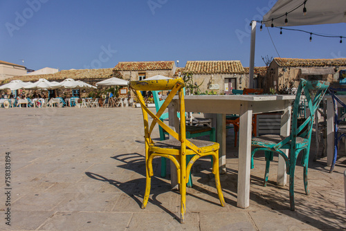  A table and two chairs in retro style, a typical Italian outdoor restaurant, a square in a small town, a yellow chair in the foreground, Marzamemi, Sicily, Italy