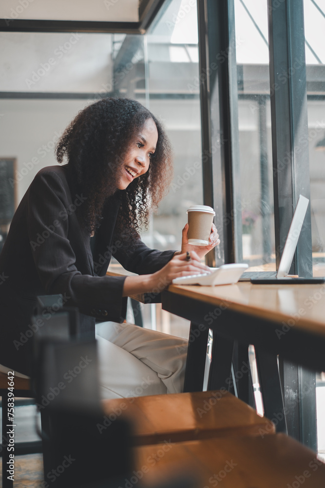 Smiling business woman calculating expenses and hand holding coffee mug, using calculator, sitting at desk with laptop and financial documents. Smiling business woman calculating expenses and hand holding coffee mug, using calculator, sitting at desk with laptop and financial documents.