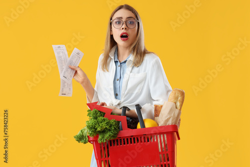 Shocked young woman with shopping basket of food and receipts on yellow background. Price rise concept photo