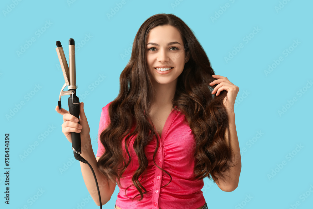 Beautiful young woman with curling iron on blue background