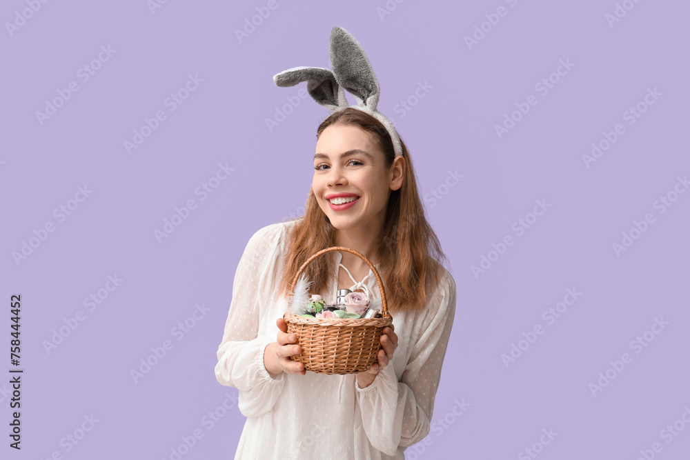 Happy smiling young woman in bunny ears headband holding basket with makeup products, flowers and Easter decor on lilac background - obrazy, fototapety, plakaty 