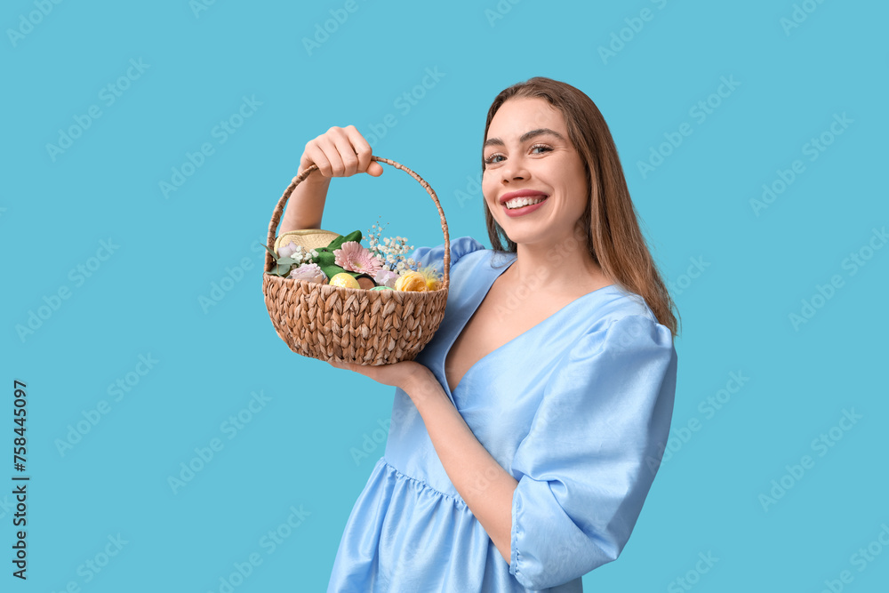 Happy smiling young woman holding basket with makeup products, flowers and Easter decor on blue background - obrazy, fototapety, plakaty 